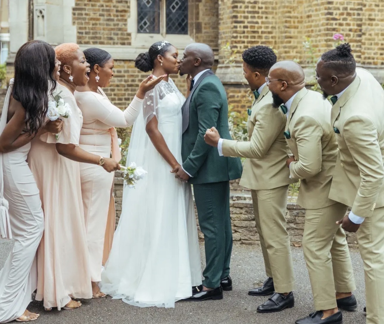 Bride and groom kissing surrounded by their cheering wedding party outside a London church