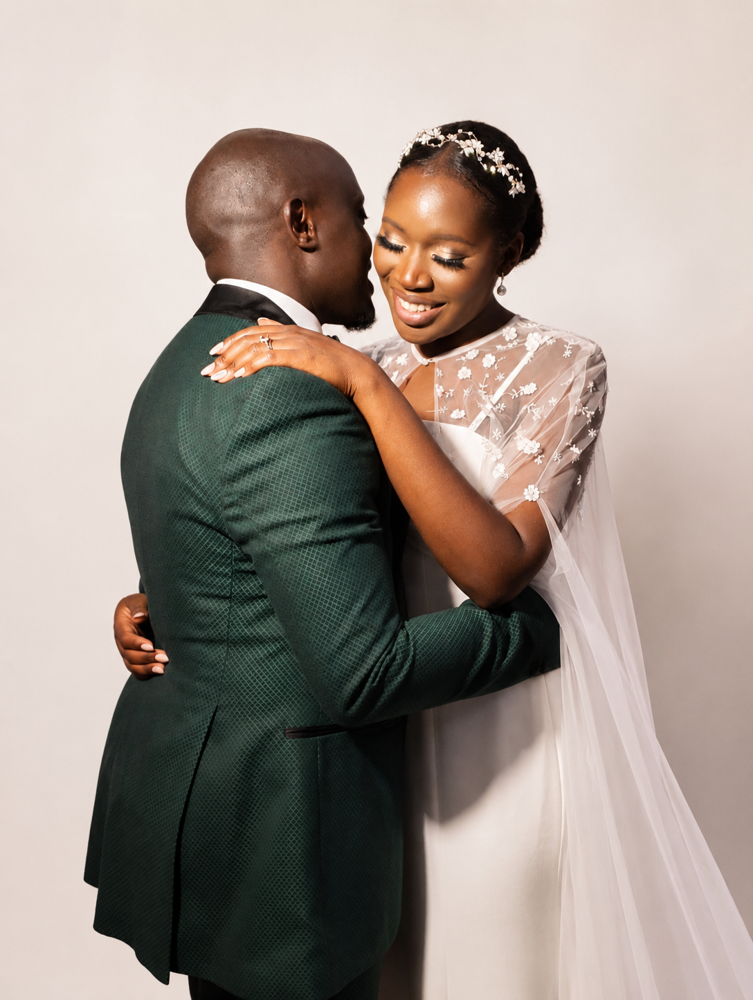 Bride in an embroidered tulle cape embracing her groom in a green tuxedo